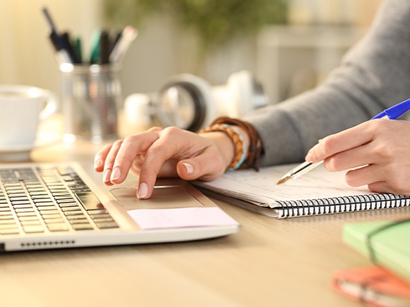 person taking notes using pen and notebook while using a laptop 